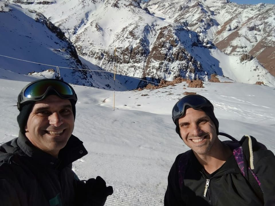 Gustavo e amigo de gorro e óculos de esqui na pista, paredões nevados da cordilheira ao fundo