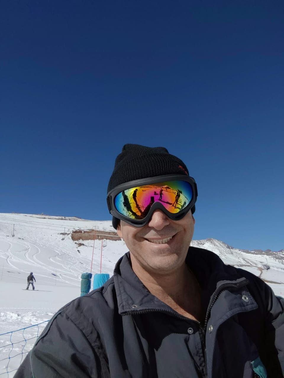 Selfie do Gustavo com óculos de esqui espelhados refletindo a pista, céu azul e encosta nevada atrás