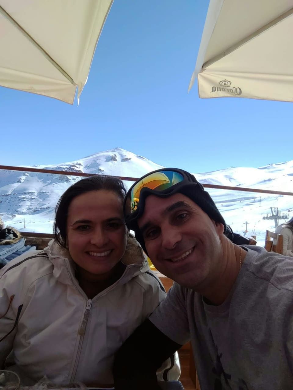 Casal sorrindo no deck de um restaurante na montanha, guarda-sóis e pico nevado do Valle Nevado ao fundo