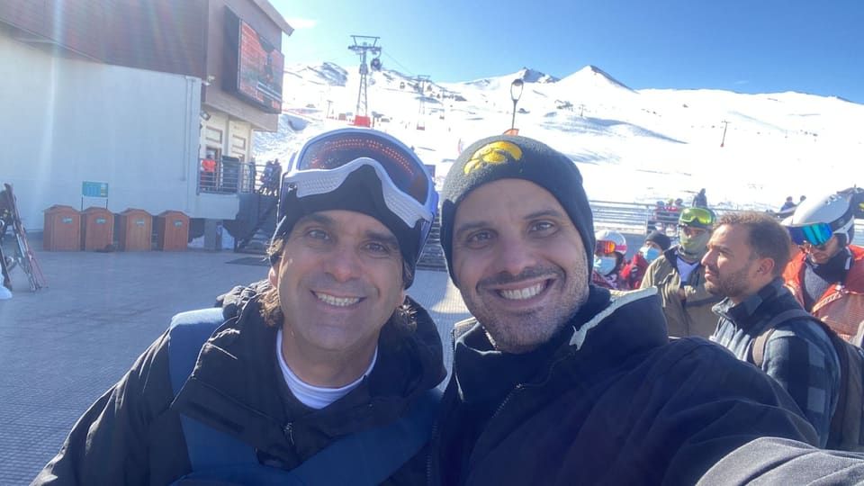 Gustavo e amigo de gorro e óculos de esqui na base do Valle Nevado, teleférico e pistas nevadas ao fundo
