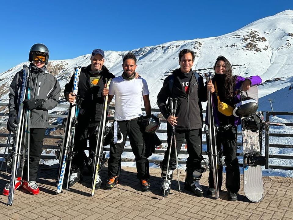 Grupo de cinco amigos com esquis e prancha de snowboard posando na neve do Valle Nevado, céu azul e montanha ao fundo