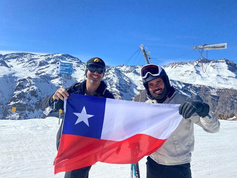 Dois amigos abrindo a bandeira do Chile na pista, ao lado das placas Fox Trot e Retorno Bajo, cordilheira nevada atrás