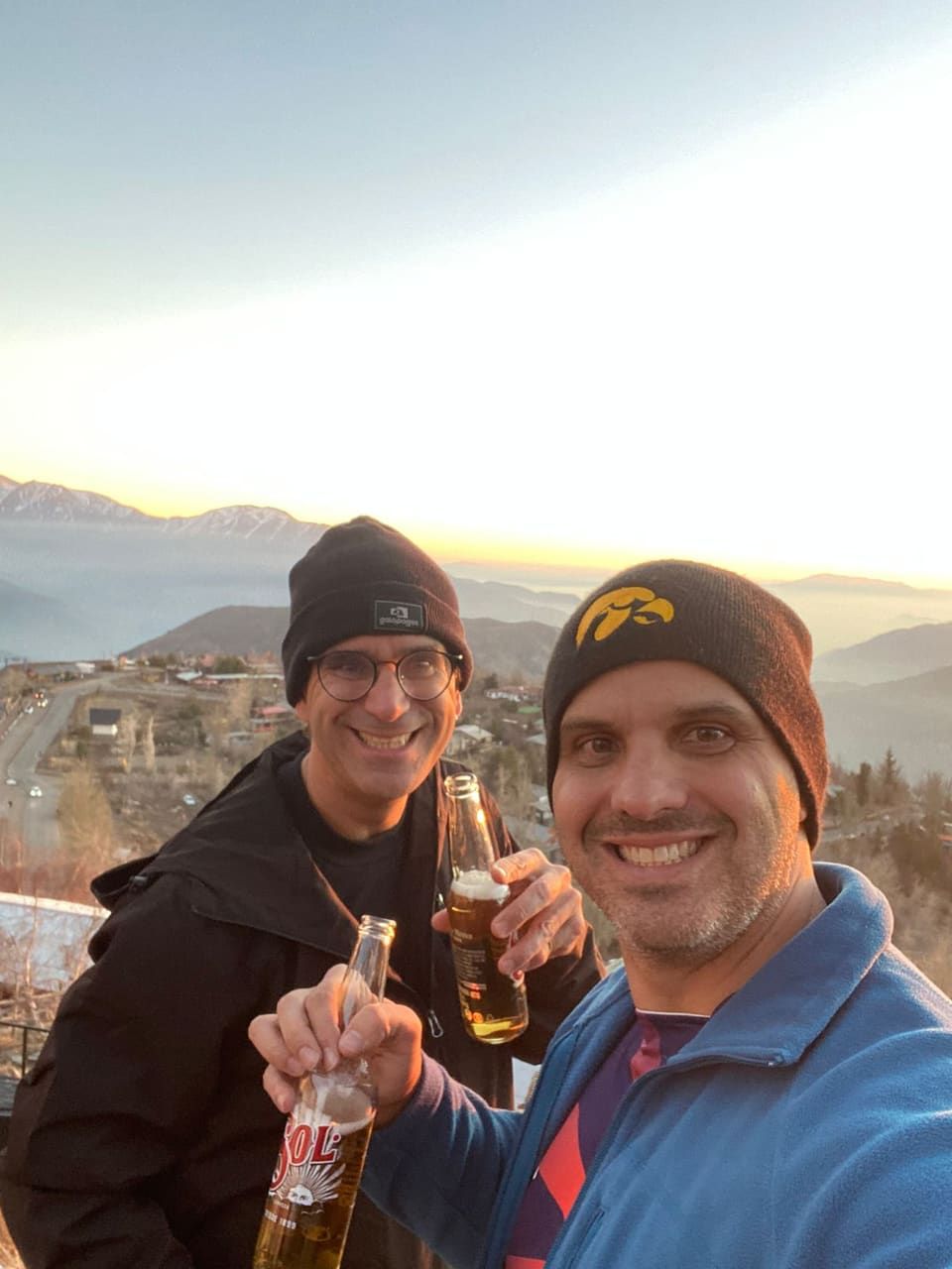 Gustavo e amigo brindando cerveja ao pôr do sol num mirante da montanha, vale e cordilheira ao fundo