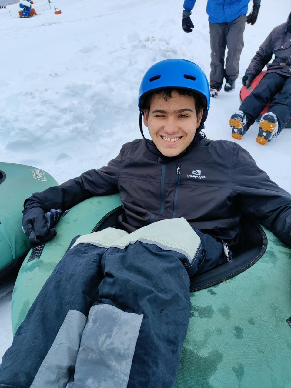 Jovem sorrindo de capacete azul numa boia de snow tubing, neve por todos os lados