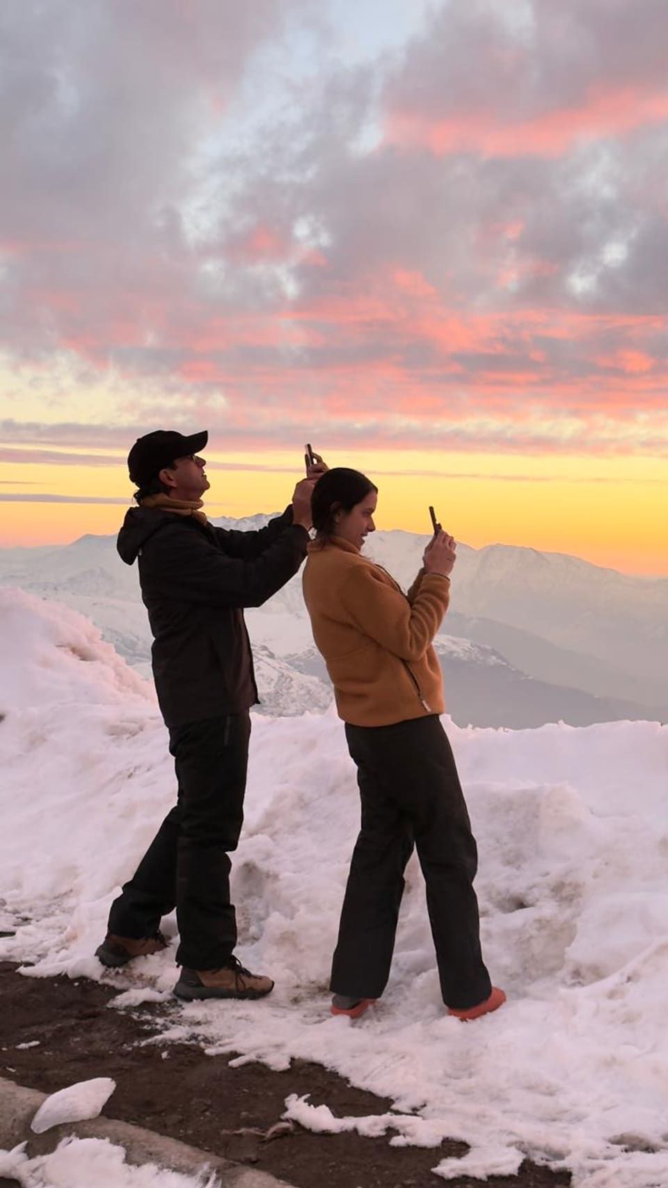 Dois viajantes fotografando com o celular o pôr do sol rosa sobre a cordilheira nevada dos Andes