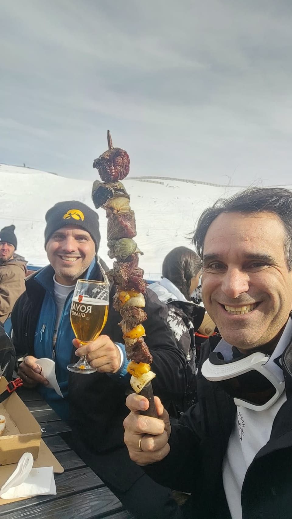 Gustavo e amigo com espetinho gigante e taça de cerveja no restaurante da pista, encosta nevada ao fundo